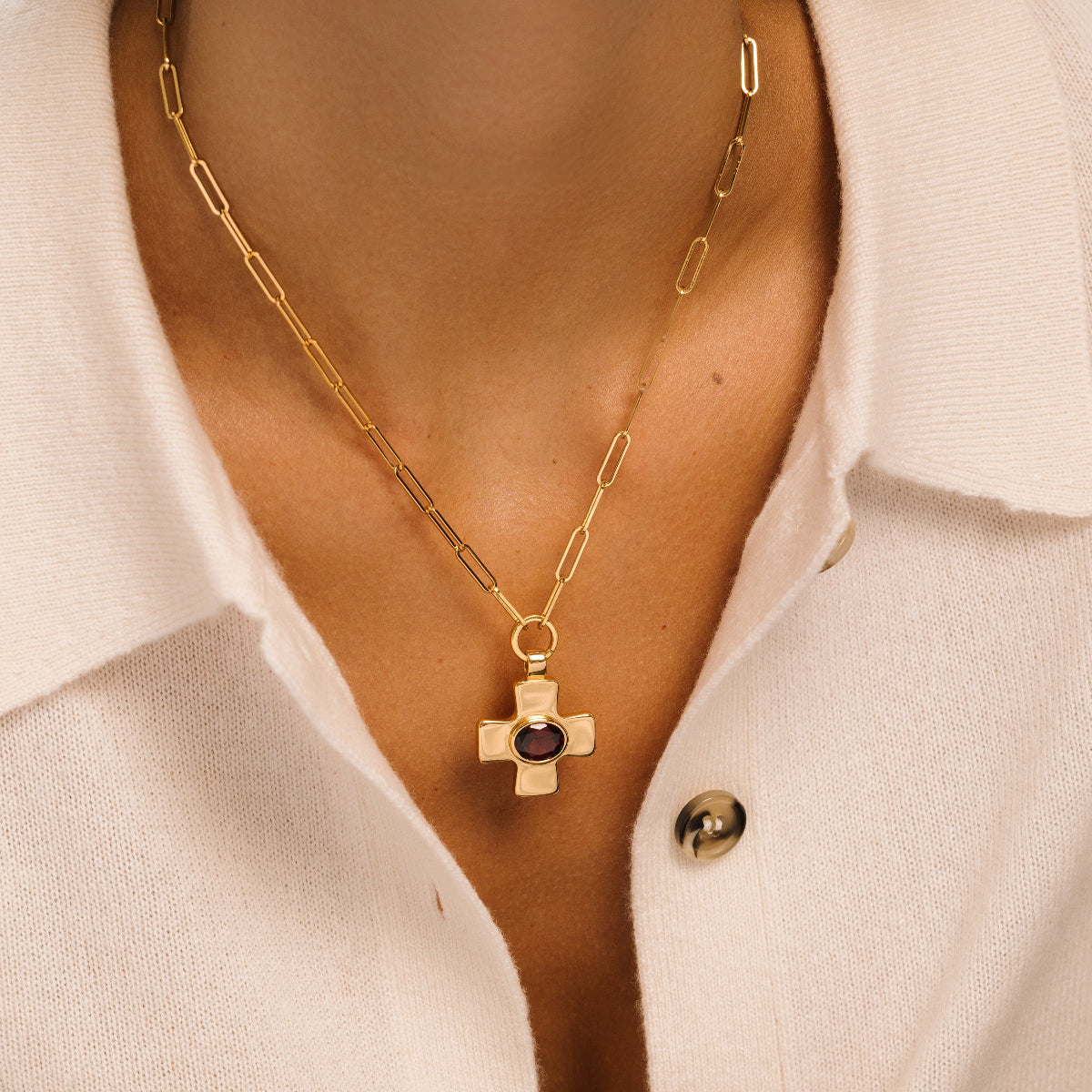 A gold paperclip chain necklace with a cross pendant featuring a dark oval stone, worn by a model in a white collared shirt.