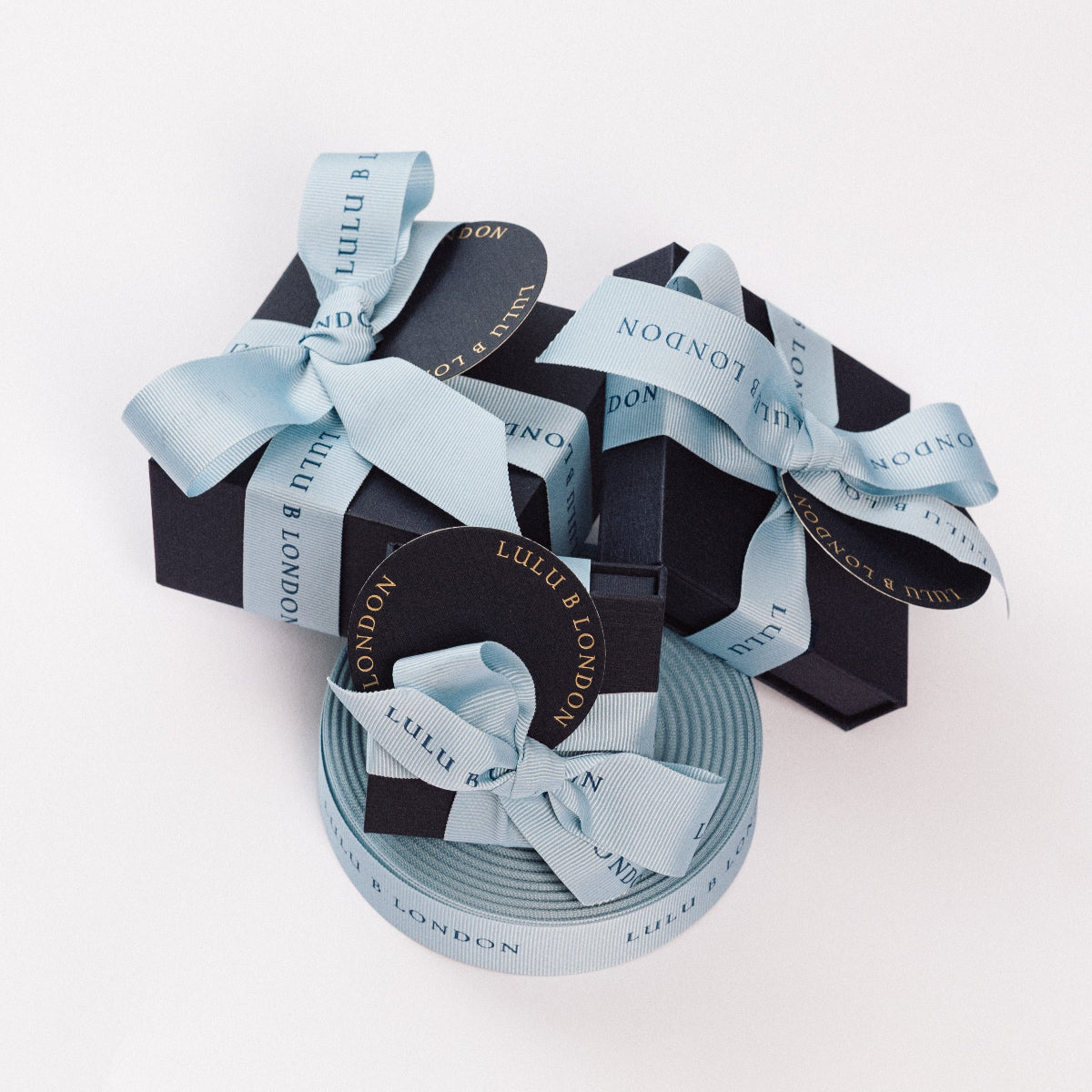 A roll of light blue ribbon with 'LULU B LONDON' printed on it, shown alongside two black gift boxes tied with matching ribbon bows on a clean white background.