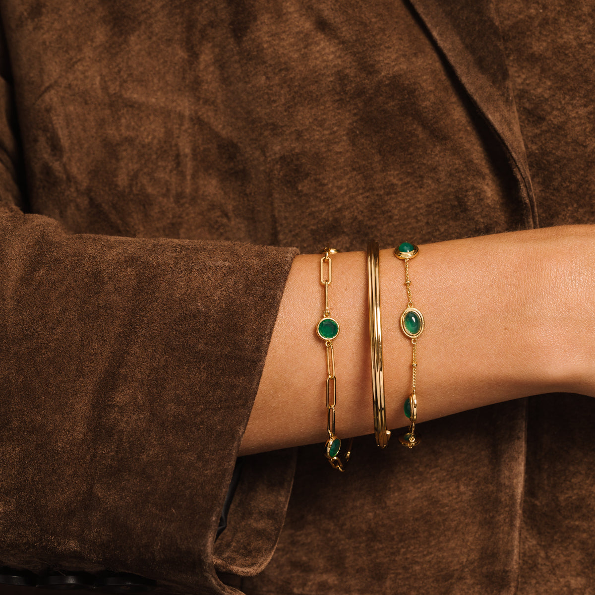 A close-up of a person's wrist wearing several gold-toned bracelets, including two with round green stones, on a brown suede jacket.