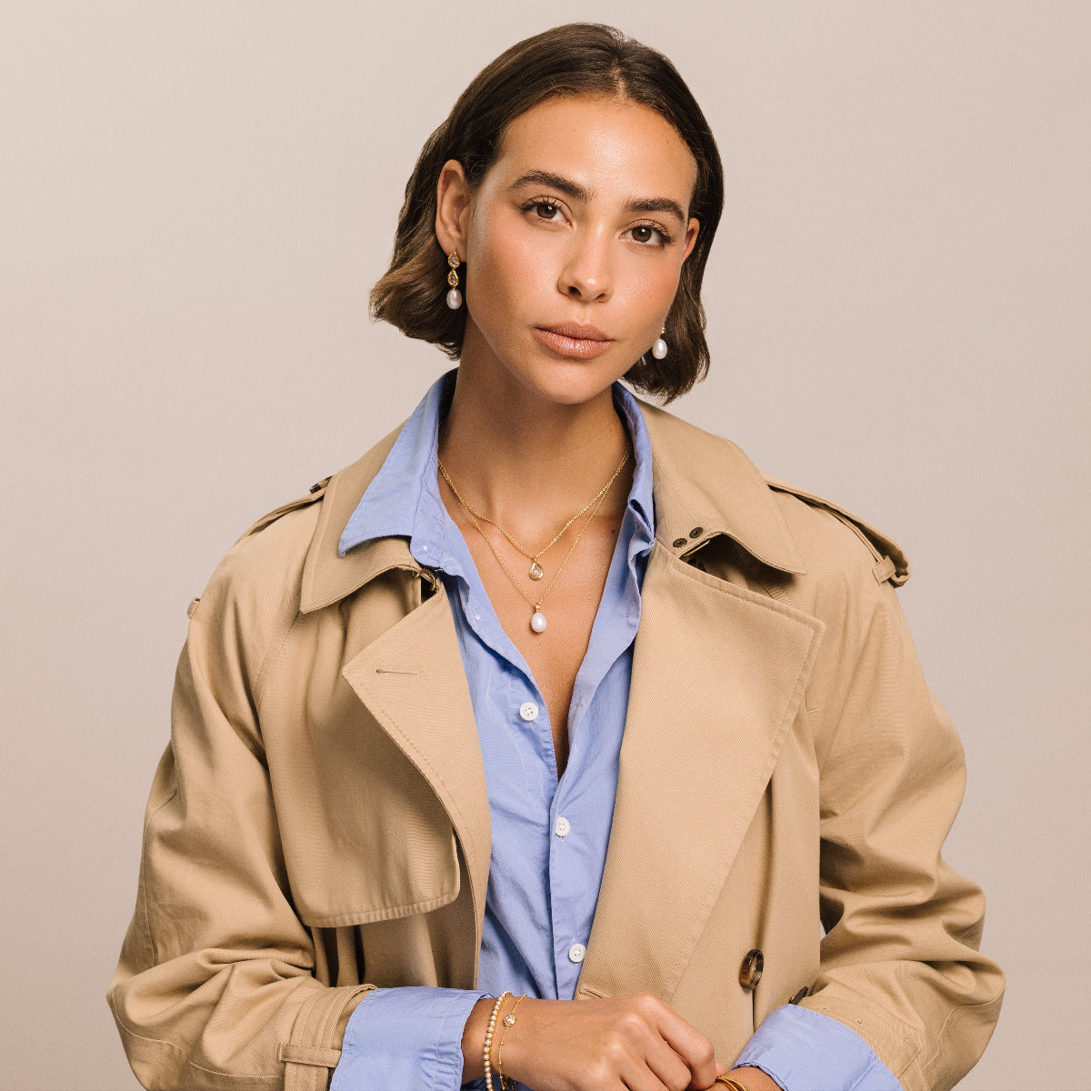 A woman wearing a beige trench coat over a light blue button-down shirt, photographed against a plain light beige studio background.